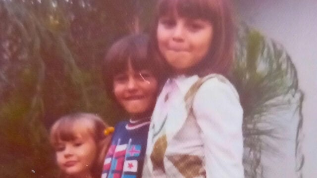 Three young children stand close together outdoors, smiling at the camera with a sense of confidence. The child on the right wears a white outfit, the middle child has a dark shirt, and the child on the left has blonde hair with bangs—a classic scene reminiscent of 70s kids. Greenery is visible behind them.