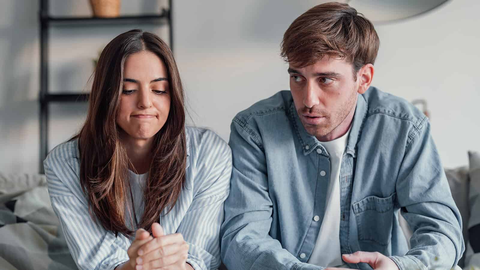 A woman with long brown hair looks down with a frustrated expression while a man beside her talks seriously, highlighting the challenges people often face in relationships. Both are sitting indoors on a couch, wearing blue shirts.