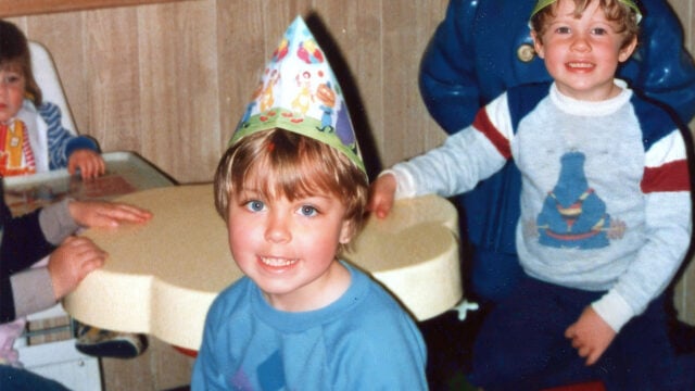 Two young children at a birthday party bring to mind the carefree spirit of 80s kids. One child wears a party hat and smiles at the camera, while the other sits nearby, also smiling. There are more kids and a yellow table in the background, capturing a joyful moment before smartphones became part of every gathering.