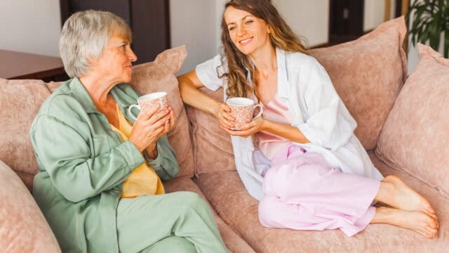An older woman and a younger woman sit on a couch, smiling and holding mugs, appearing to enjoy a friendly conversation together.