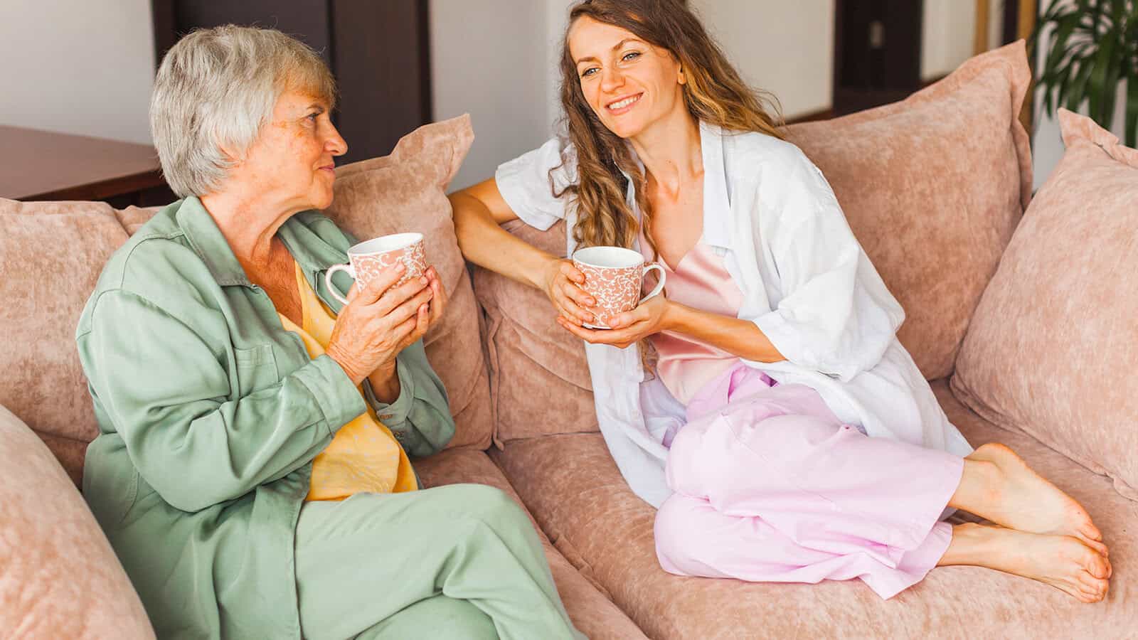 An older woman and a younger woman sit on a couch, smiling and holding mugs, appearing to enjoy a friendly conversation together.
