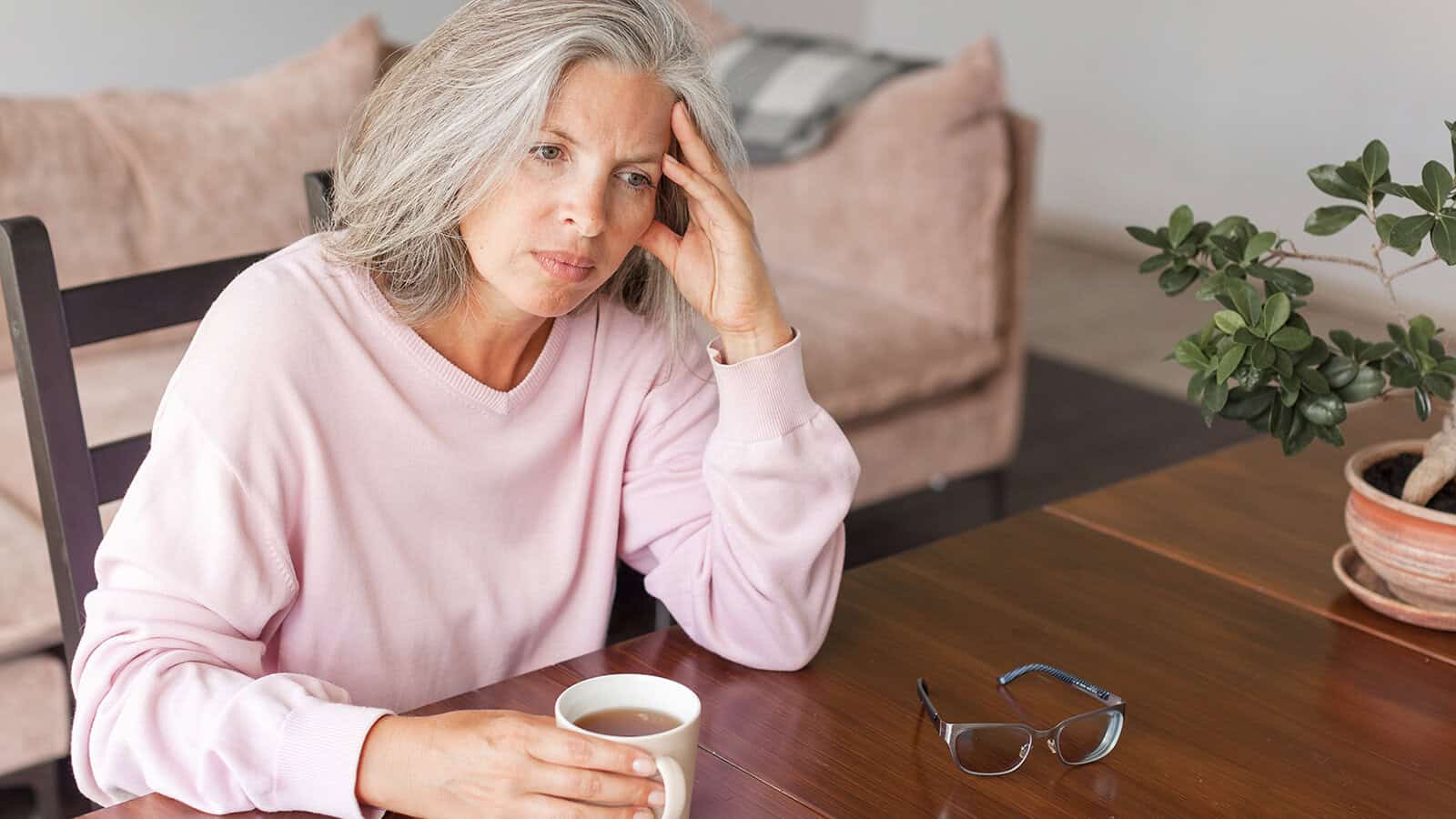A woman with gray hair sits at a table, holding a cup and resting her head on her hand, looking thoughtful or worried. Glasses and a potted plant are on the table. A sofa is in the background.