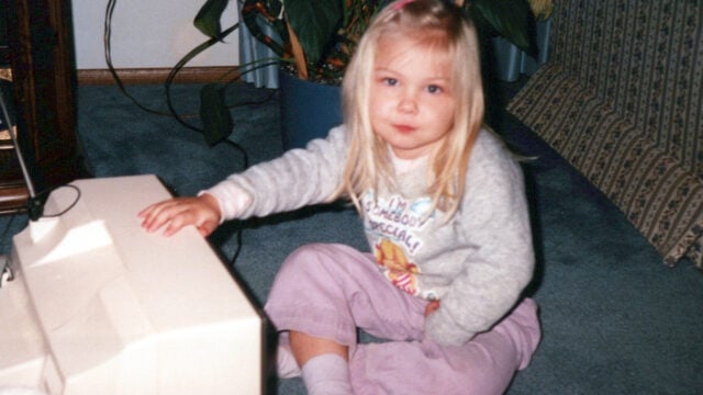 A young girl with long blonde hair sits on a blue carpet, wearing a gray sweater and purple pants, with one hand resting on a beige computer monitor. A large plant and patterned furniture are in the background.