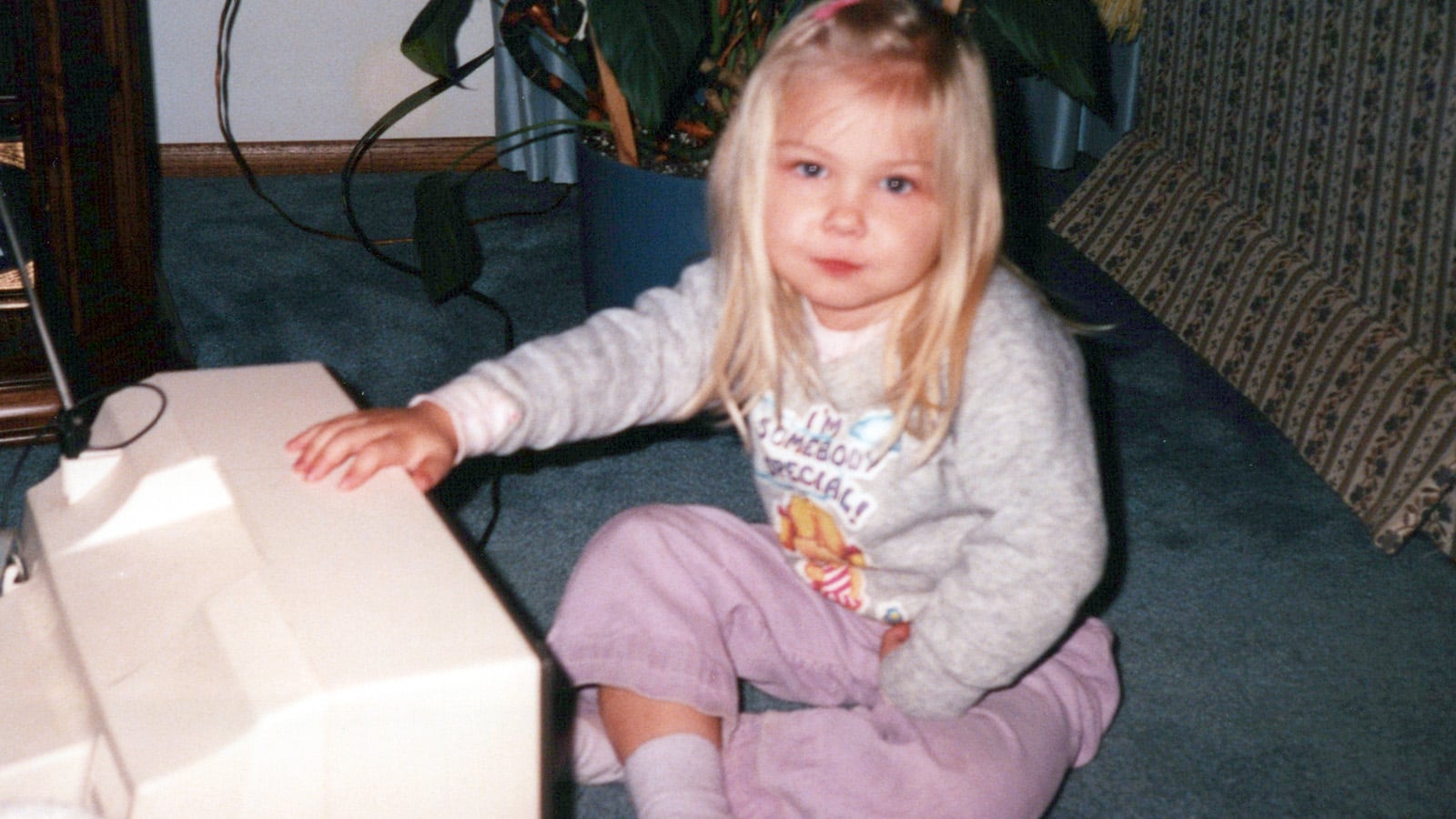 A young girl with long blonde hair sits on a blue carpet, wearing a gray sweater and purple pants, with one hand resting on a beige computer monitor. A large plant and patterned furniture are in the background.