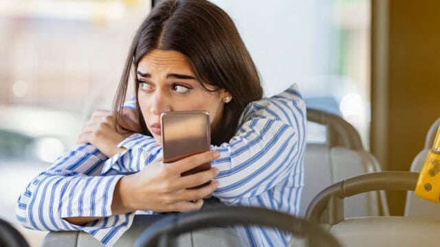 A woman with long hair and a striped shirt sits on a bus, hugging her knees and holding a phone, looking worried and gazing out the window.