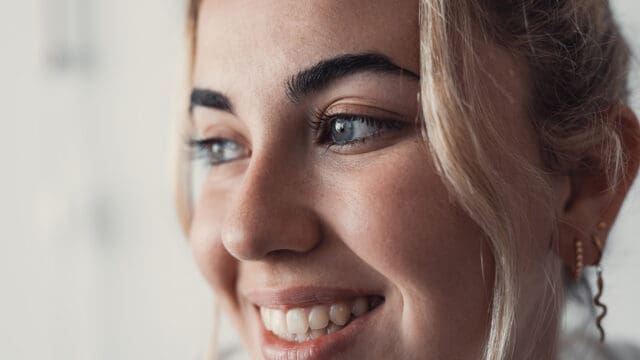 A close-up of a smiling person with light skin, blue eyes, and blonde hair. The image highlights their clear skin, white teeth, and gold earrings, with soft natural lighting coming from the left.