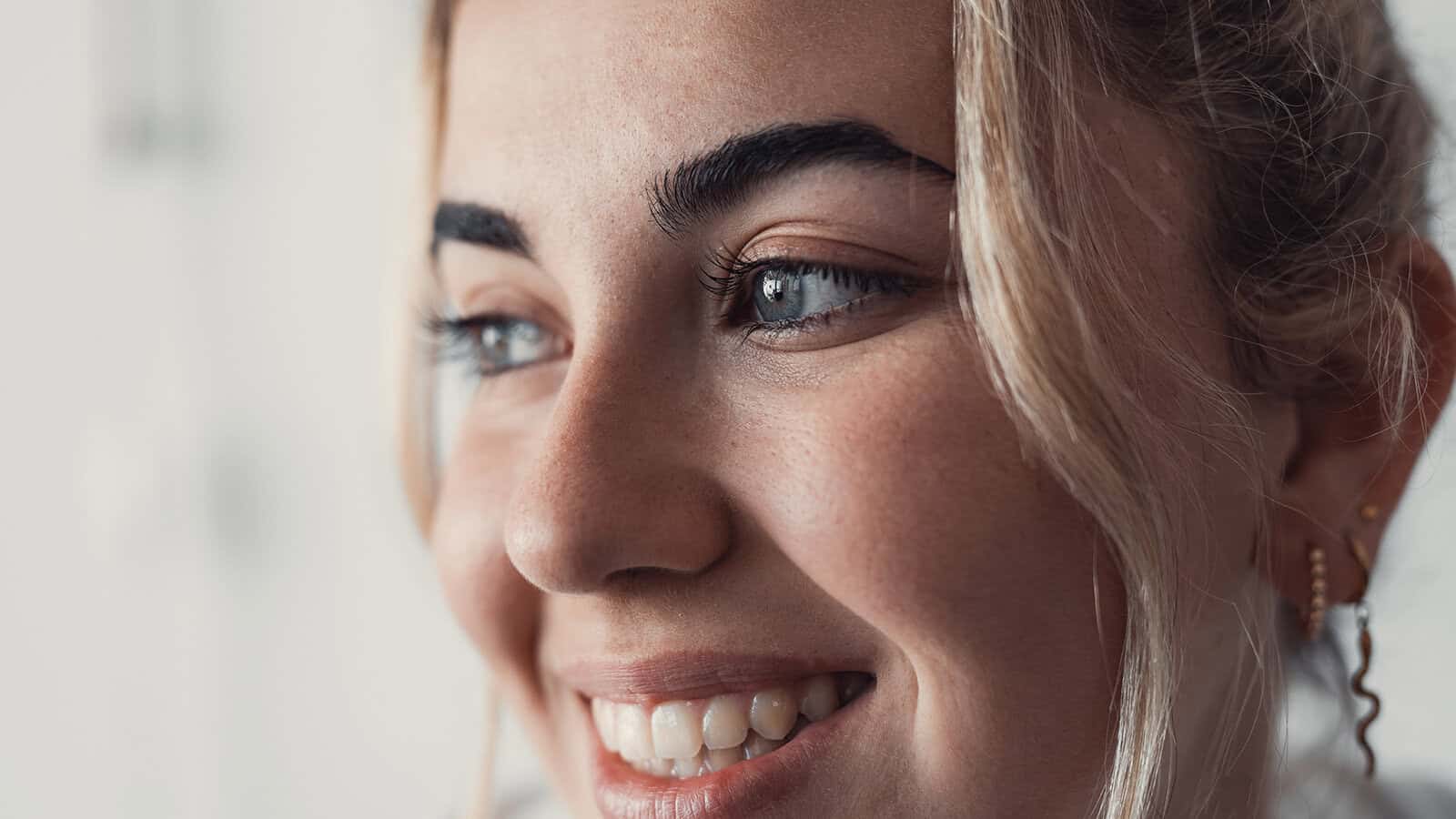 A close-up of a smiling person with light skin, blue eyes, and blonde hair. The image highlights their clear skin, white teeth, and gold earrings, with soft natural lighting coming from the left.