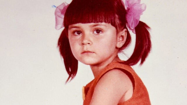 Young girl with straight brown hair in pigtails, each tied with a large pink bow, wearing an orange sleeveless top, looking slightly to the side with a neutral expression against a plain background—a subtle nod to the classic styles favored by people raised in the 60s and 70s.