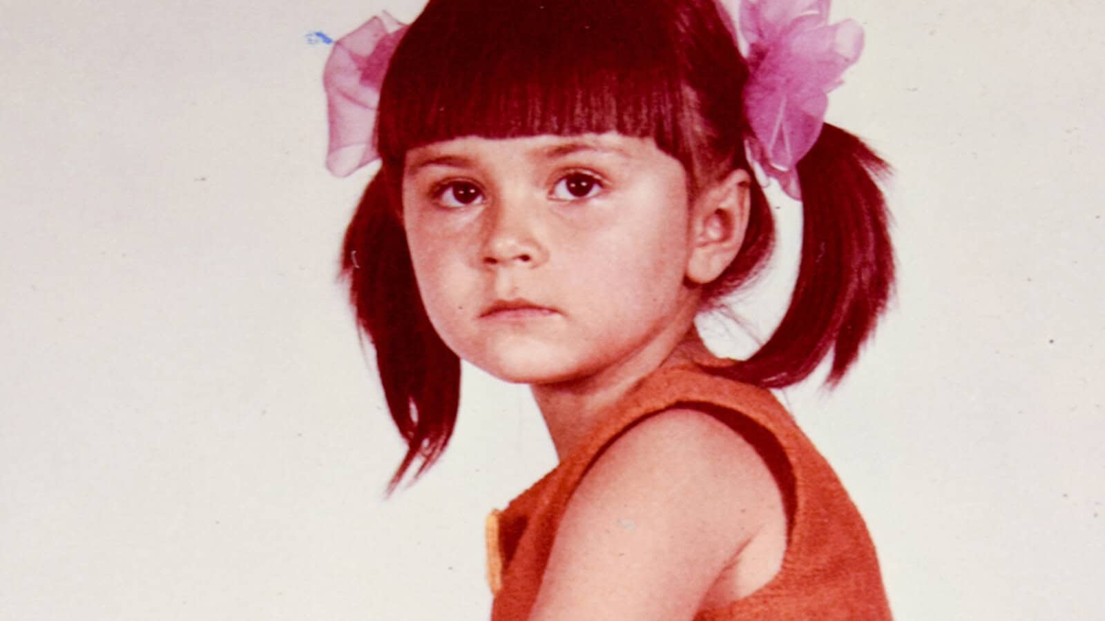 Young girl with straight brown hair in pigtails, each tied with a large pink bow, wearing an orange sleeveless top, looking slightly to the side with a neutral expression against a plain background—a subtle nod to the classic styles favored by people raised in the 60s and 70s.