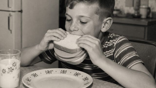 A young boy in a striped shirt eats a sandwich at a table with an empty plate and a glass of milk. The photo is in black and white, giving it a vintage feel that echoes the vintage eating habits and retro foods of the 60s and 70s.