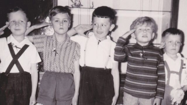 Five young boys stand in a row inside a room, saluting. Four wear suspenders and shorts, while one wears a striped sweater. The photo is black and white, with a cabinet visible in the background.