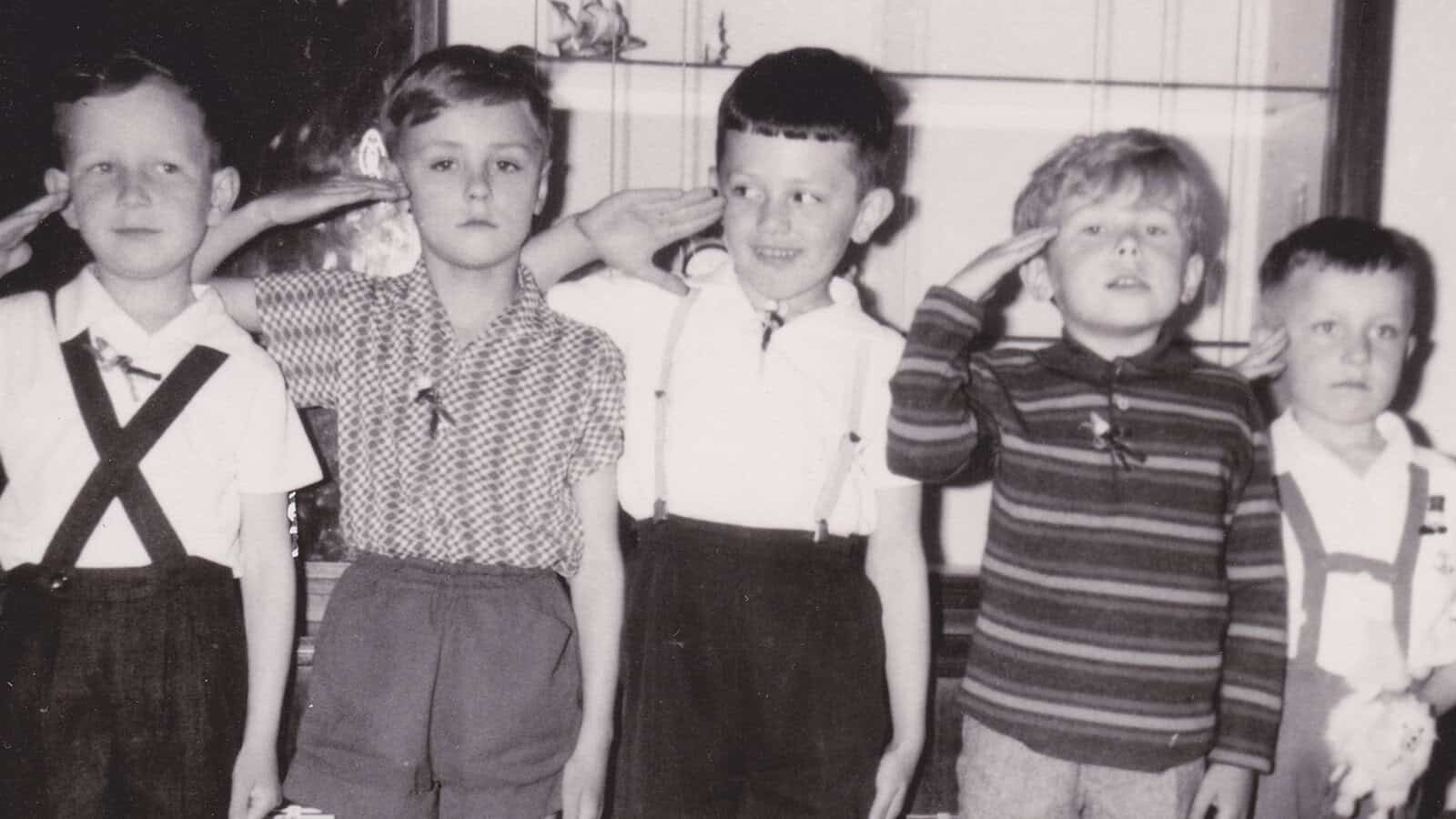 Five young boys stand in a row inside a room, saluting. Four wear suspenders and shorts, while one wears a striped sweater. The photo is black and white, with a cabinet visible in the background.