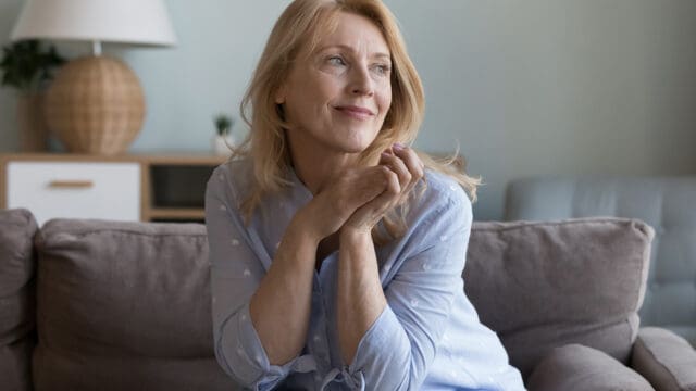 A woman with blonde hair sits on a gray couch, wearing a light blue shirt, smiling softly and looking off to the side. There is a lamp and a small plant in the background.