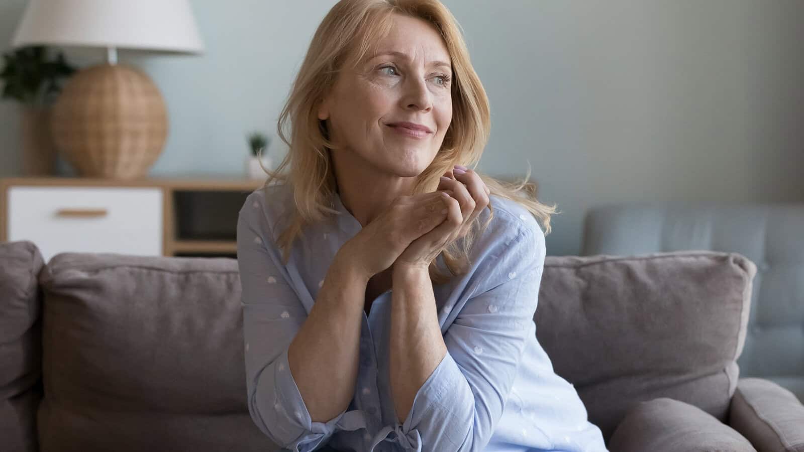 A woman with blonde hair sits on a gray couch, wearing a light blue shirt, smiling softly and looking off to the side. There is a lamp and a small plant in the background.