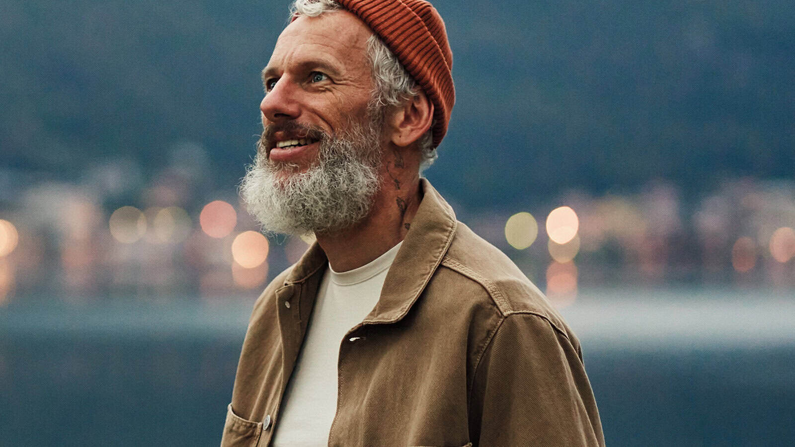 A smiling man with a gray beard wearing an orange beanie, tan jacket, and white shirt stands outdoors near water with blurred city lights in the background.
