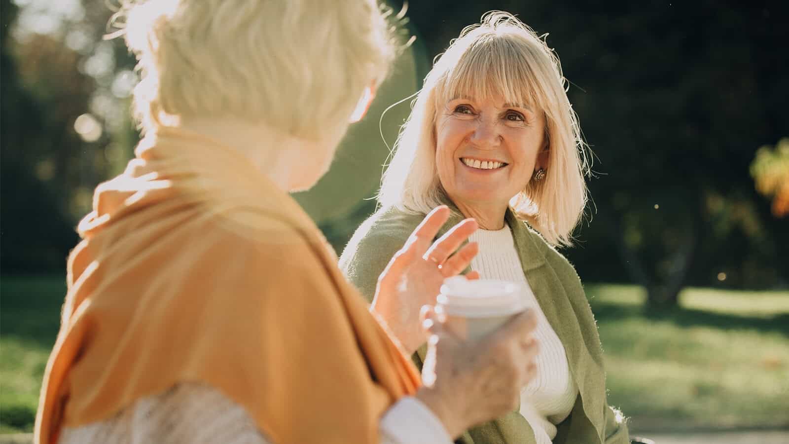 Two older women sit outside, smiling and talking. One wears a green sweater and holds a coffee cup, while the other wears an orange scarf. Sunlight shines in the background, creating a warm and friendly atmosphere.