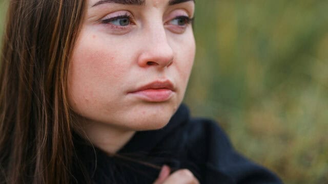 A woman with long brown hair looks off to the side with a serious, contemplative expression. She is wearing a black top and is outdoors, with a blurred green background.