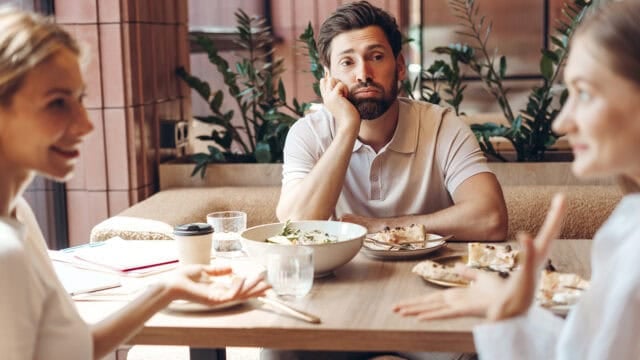 A man sits at a table with two women, looking bored while they talk. Plates of food, salads, and drinks are on the table. The setting appears to be a casual restaurant or café with plants in the background.