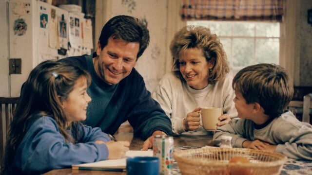 A family of four sits around a kitchen table, smiling and talking. The parents face their two young children, a girl and a boy. A basket of fruit and drinks are on the table, and a window is in the background.