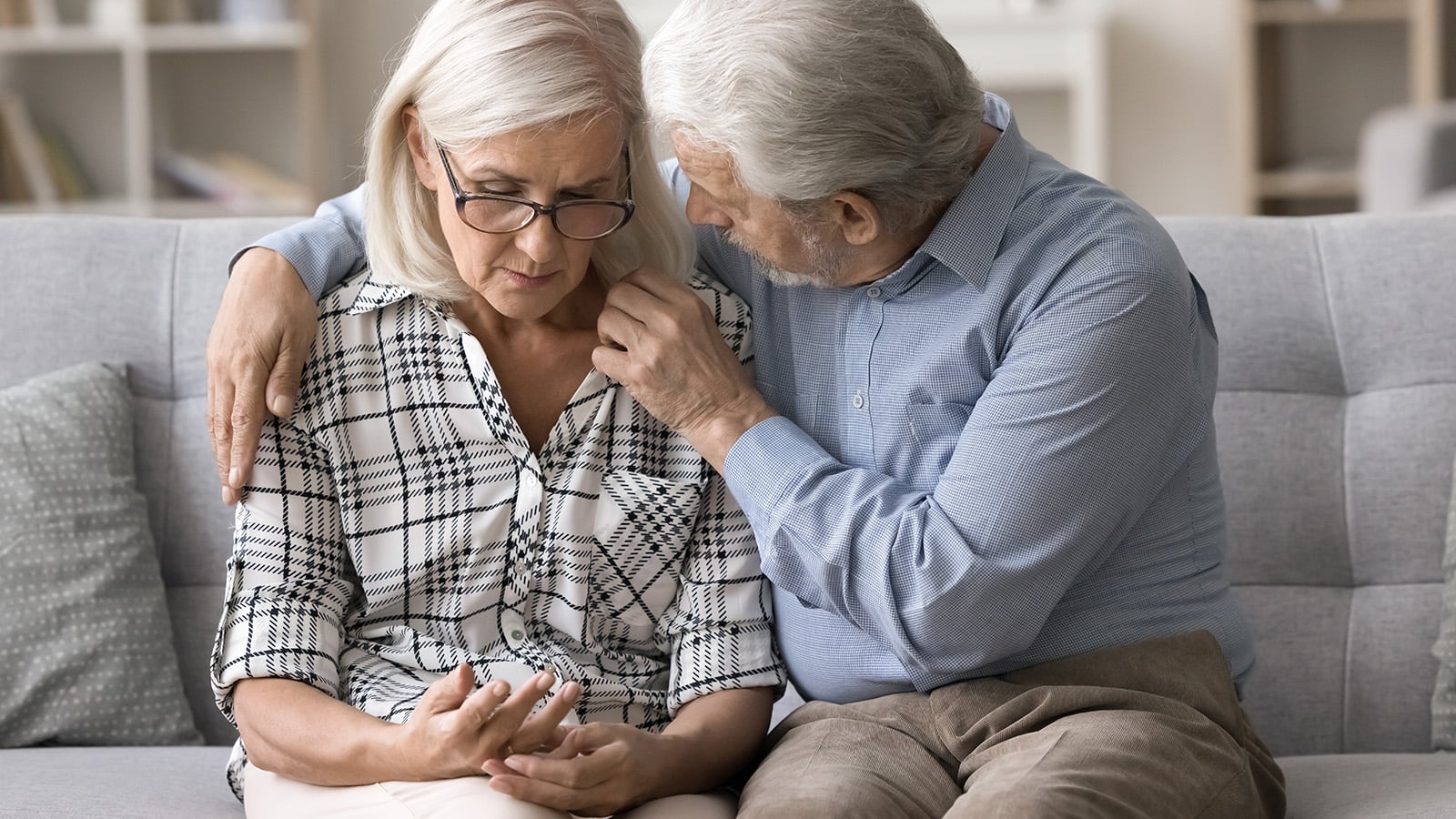 An older man comforts a sad older woman sitting next to him on a couch. She looks down at her hand, appearing upset, while he puts his arm around her and gently touches her shoulder.