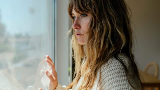 A woman with long, wavy hair and a knit sweater looks out a window with a thoughtful expression, her hand resting on the glass. Sunlight softly lights her face.