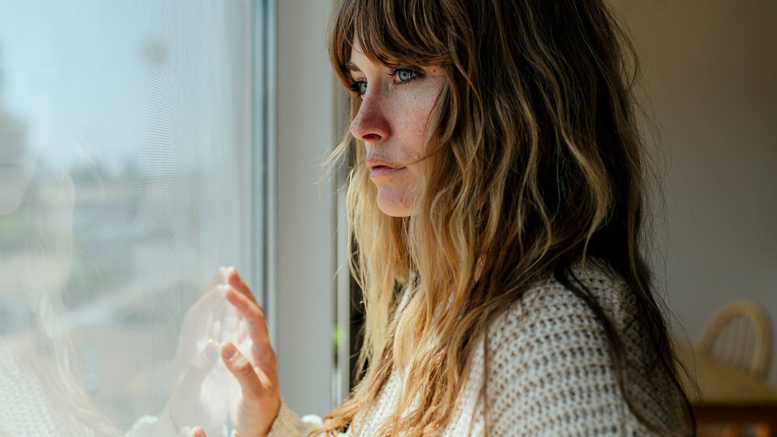A woman with long, wavy hair and a knit sweater looks out a window with a thoughtful expression, her hand resting on the glass. Sunlight softly lights her face.