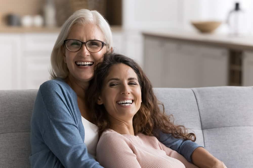 Two women—one a Gen X with long brown hair, the other an older Boomer with gray hair and glasses—sit close together on a couch, smiling and hugging in a bright, cozy living room.