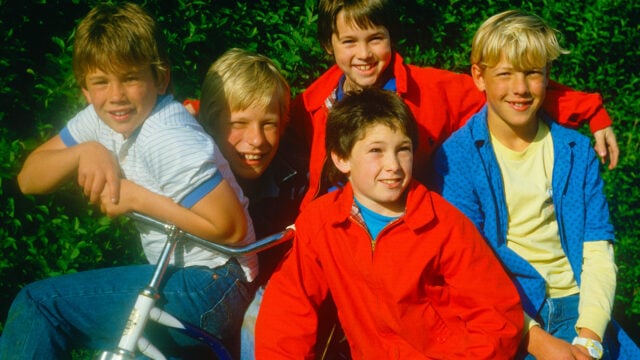 Five smiling children sit closely together outdoors, with one holding a bicycle. They wear colorful clothing, and green foliage is visible in the background.