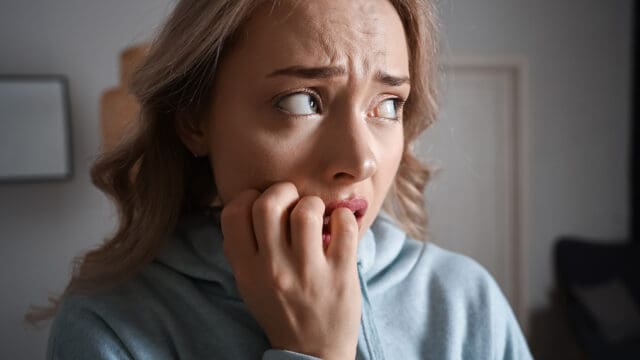 A woman with light hair and a blue hoodie looks anxious, biting her nails and glancing to the side with a worried expression.