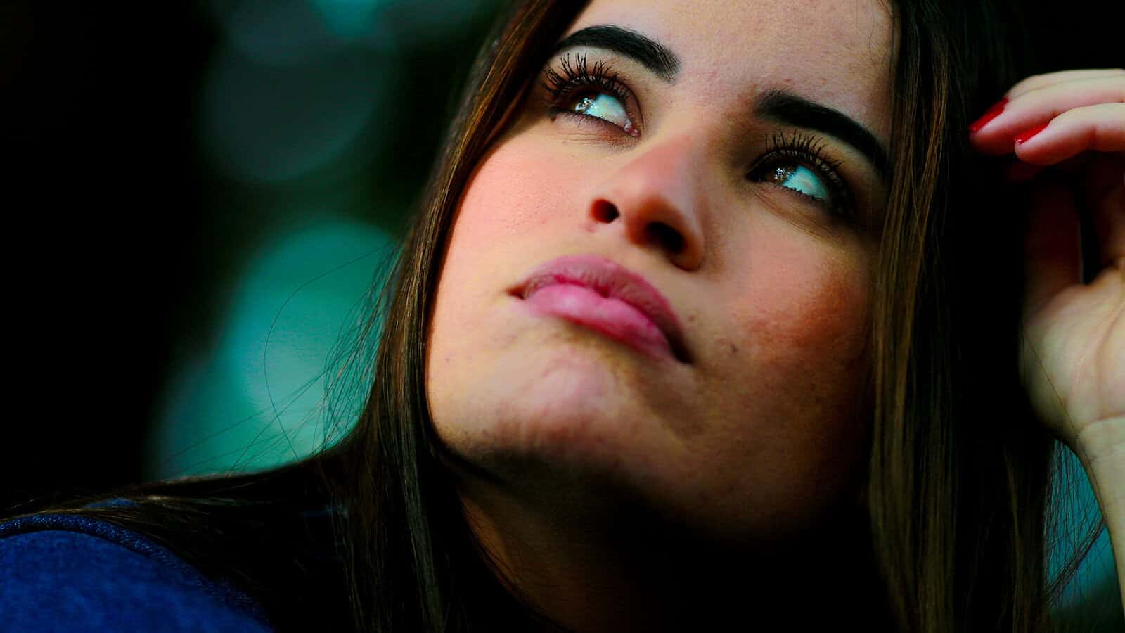 A woman with long dark hair looks upward thoughtfully, her hand near her head, with a blurred background of blue and green tones.