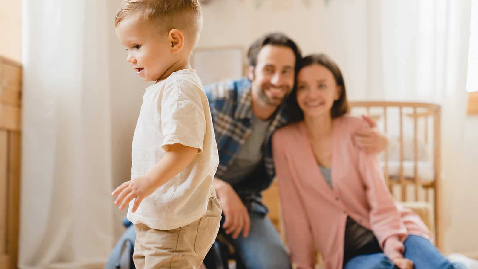 A toddler walks in the foreground, while two adults, smiling and sitting close together, are blurred in the background inside a bright, cozy room with a crib.