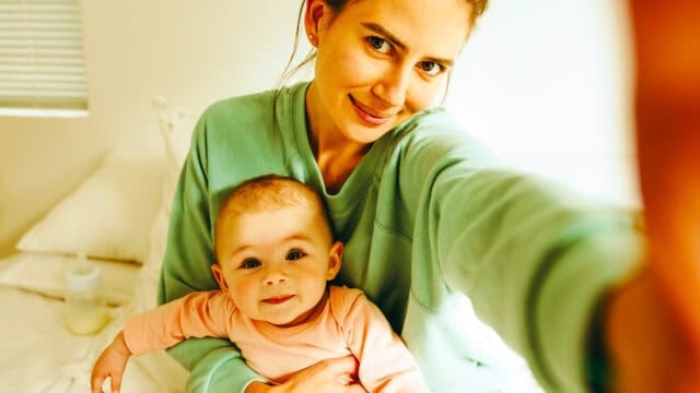 A woman in a green sweater takes a selfie while holding a smiling baby in a pink outfit. They are sitting on a bed in a bright, cozy room.