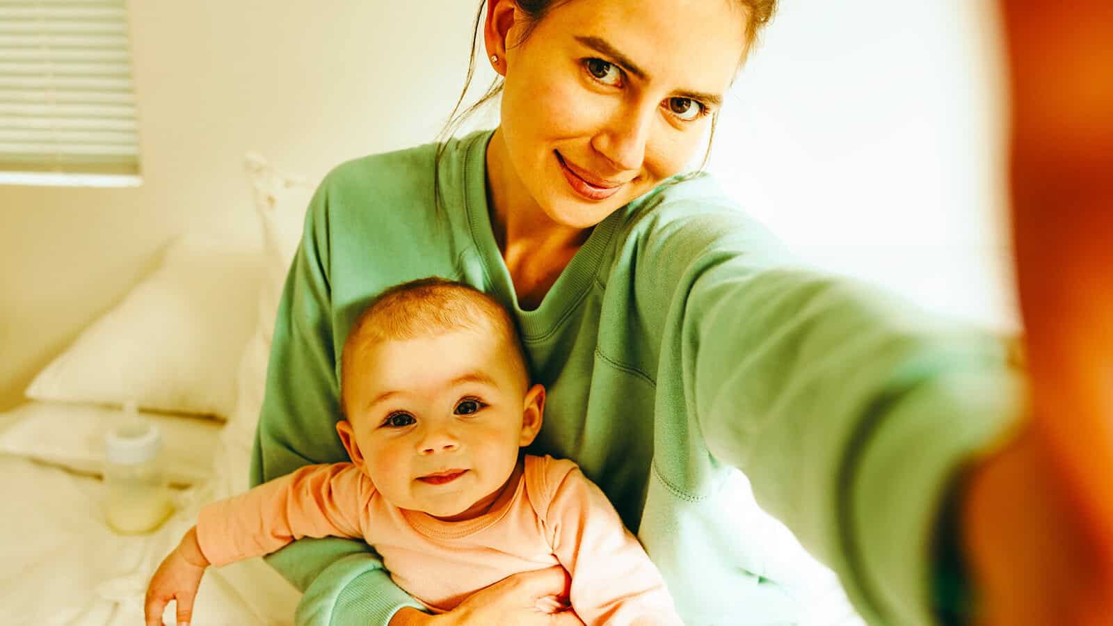 A woman in a green sweater takes a selfie while holding a smiling baby in a pink outfit. They are sitting on a bed in a bright, cozy room.