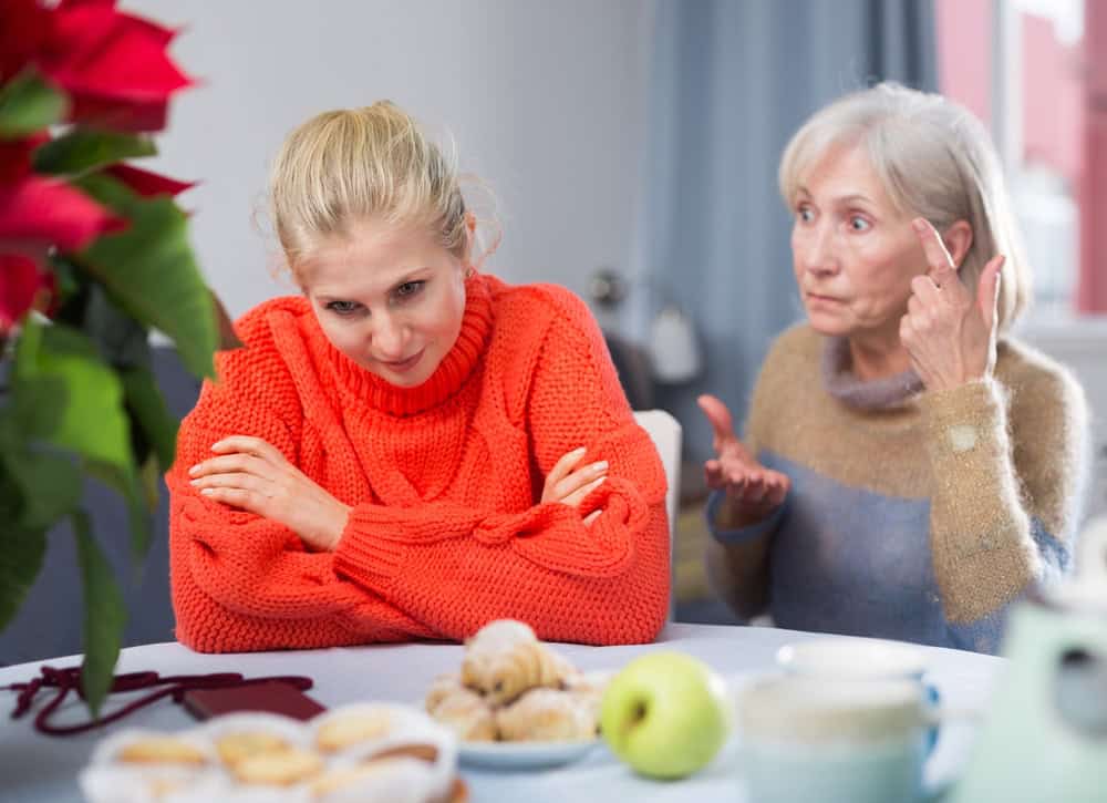 An older woman gestures and frowns at her adult child, a younger woman in an orange sweater, who looks away with arms crossed at a table with pastries and fruit.