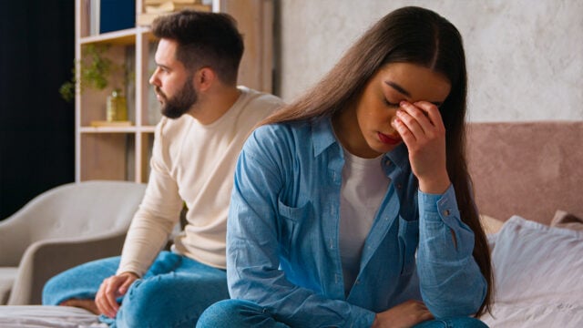 A woman sits on a bed, looking upset with her hand on her face, while a man sits behind her, turned away and gazing into the distance. Both appear distressed, suggesting a tense or emotional moment that may reflect challenges in personal growth or difficulties related to social habits.