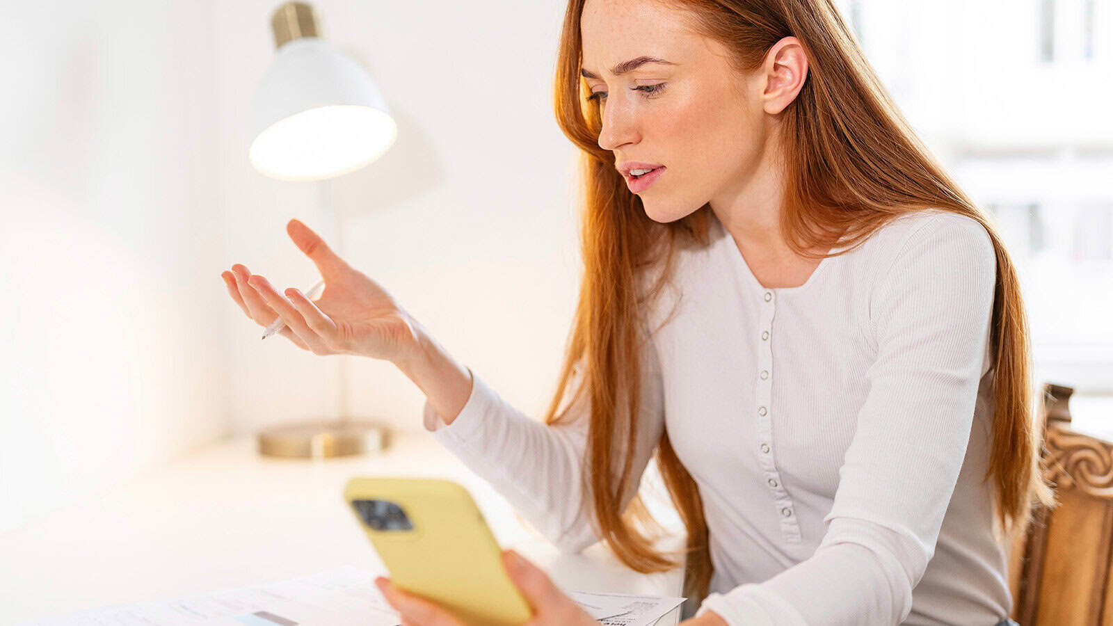 A woman with long red hair sits at a desk holding a yellow smartphone, looking frustrated or confused. Papers and a pen are on the desk, and a lamp is in the background.