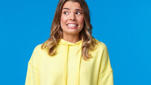 A woman in a yellow hoodie stands against a blue background, looking anxious or nervous with wide eyes and a tense, forced smile.