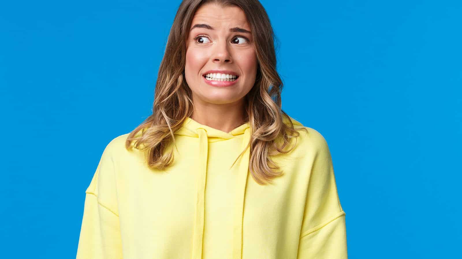 A woman in a yellow hoodie stands against a blue background, looking anxious or nervous with wide eyes and a tense, forced smile.