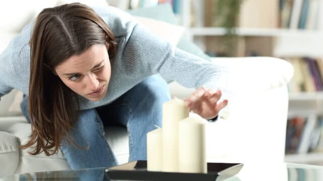 A woman intently adjusts three white candles on a black tray while sitting on a couch in a living room.