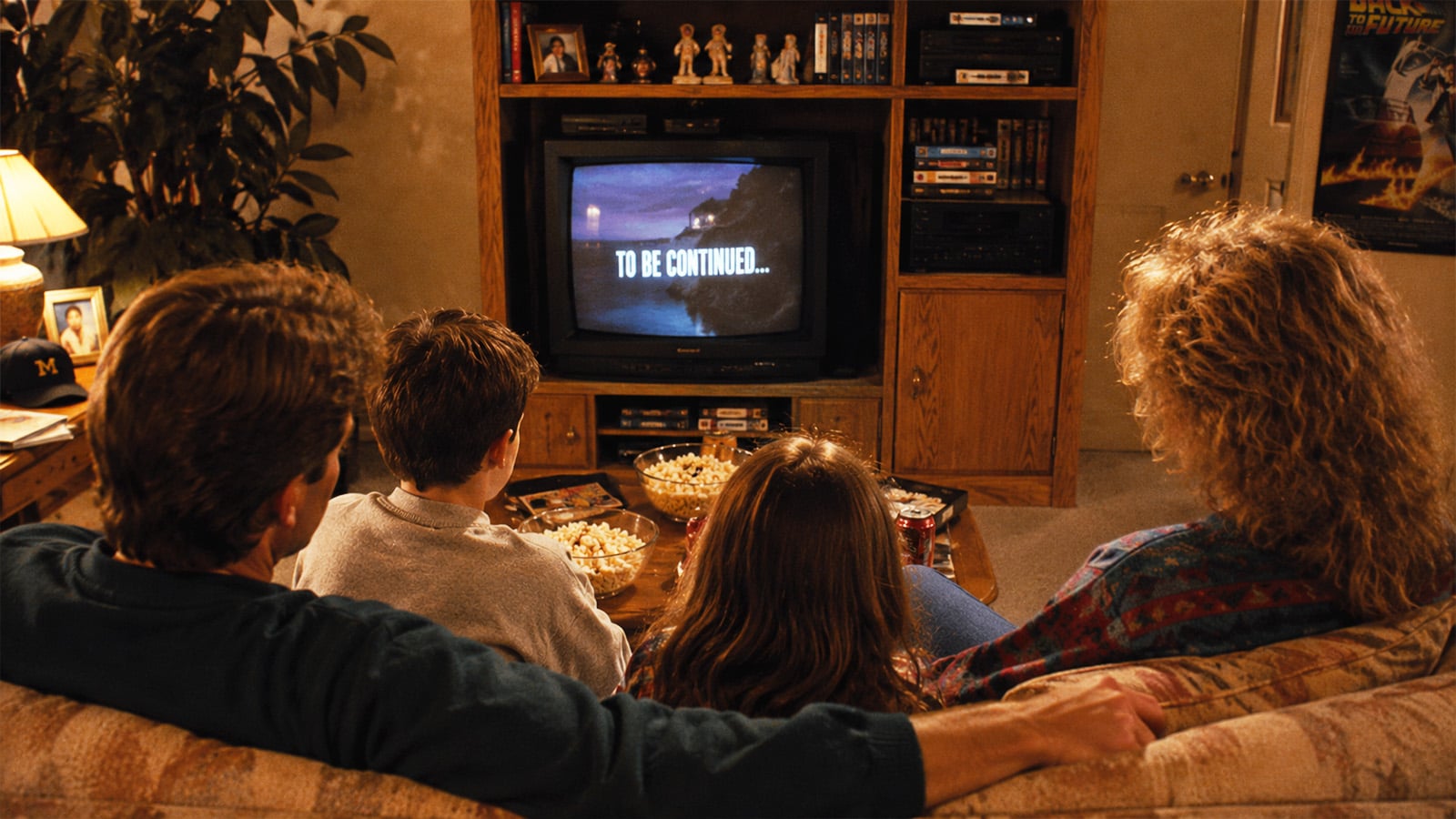 A family of four sits on a couch watching a TV that displays TO BE CONTINUED. They have popcorn bowls on the table, and the living room is warmly lit with shelves, movies, and decorations in the background.