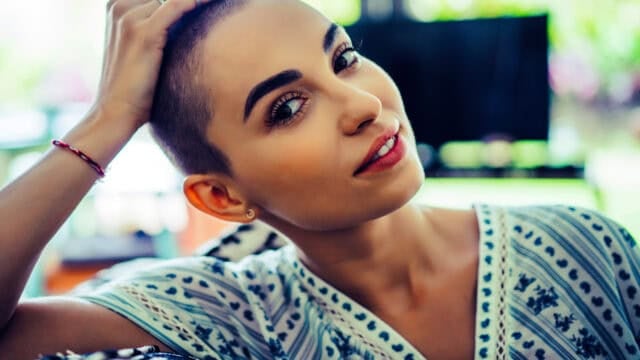 A woman with a shaved head and bold eyebrows smiles slightly, wearing a patterned shirt and touching her head, with a blurred indoor background.