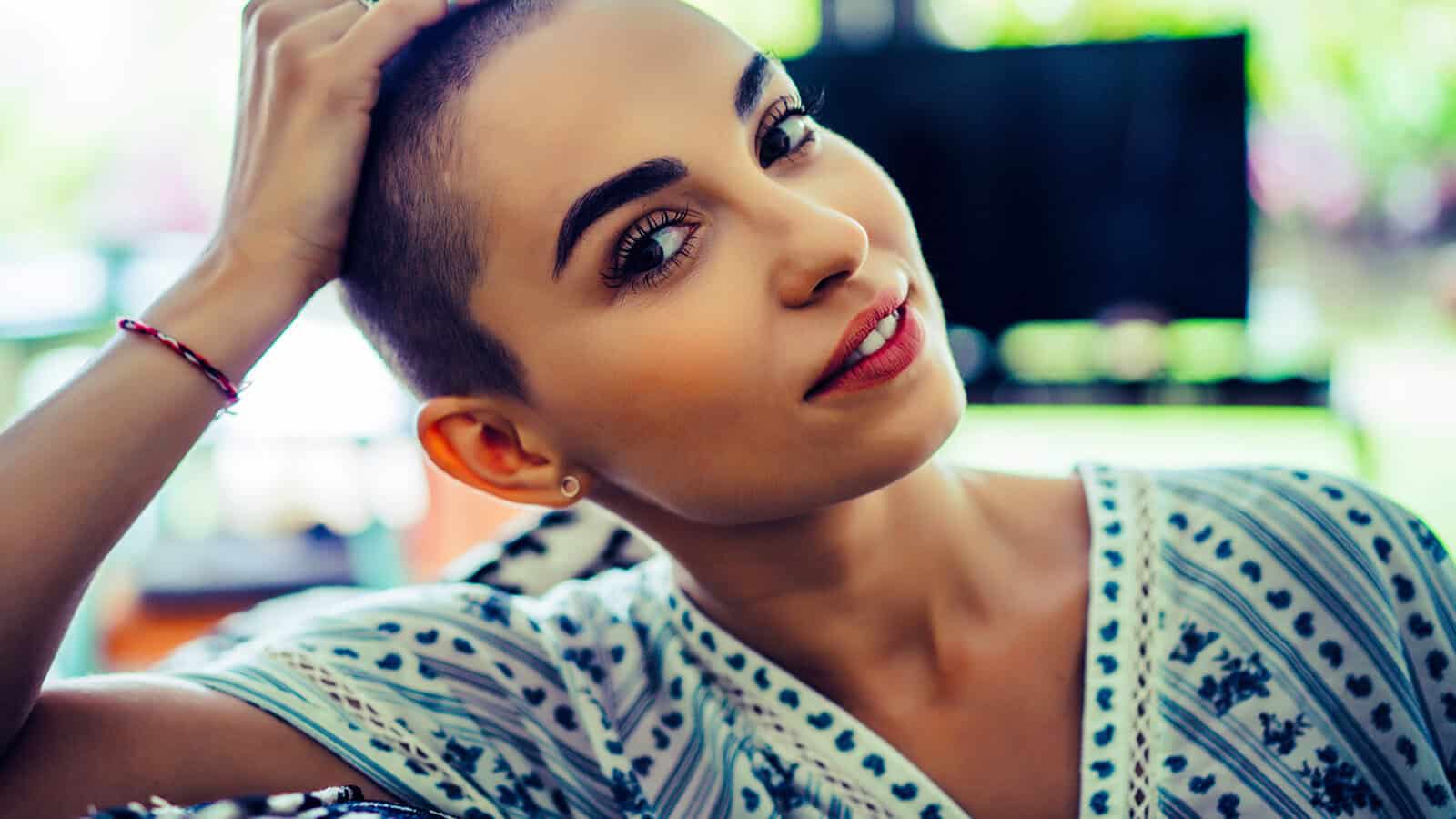 A woman with a shaved head and bold eyebrows smiles slightly, wearing a patterned shirt and touching her head, with a blurred indoor background.
