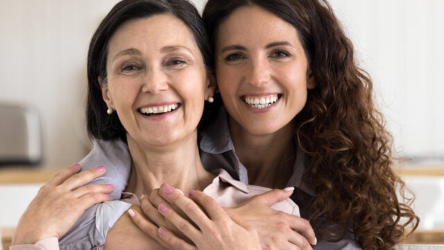 Two women smiling and hugging each other closely, sitting indoors. They both have brown hair and are wearing light-colored tops, looking happy and content.