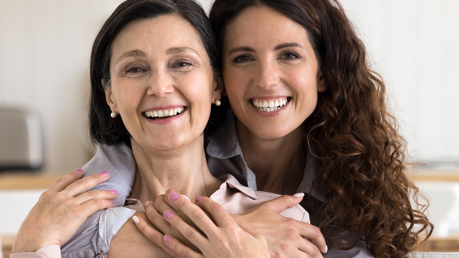 Two women smiling and hugging each other closely, sitting indoors. They both have brown hair and are wearing light-colored tops, looking happy and content.