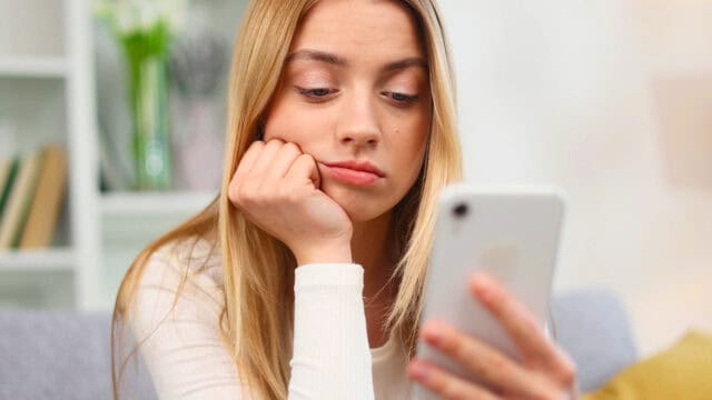 A young woman with long blonde hair rests her chin on her hand, looking bored or disappointed as she stares at a white smartphone. She is sitting indoors on a couch with blurry shelves in the background.