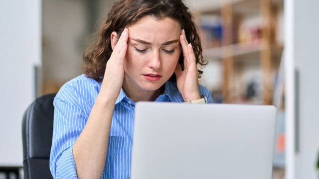 A woman sitting at a desk with her hands on her temples, looking stressed or worried while working on a laptop in a home office setting.