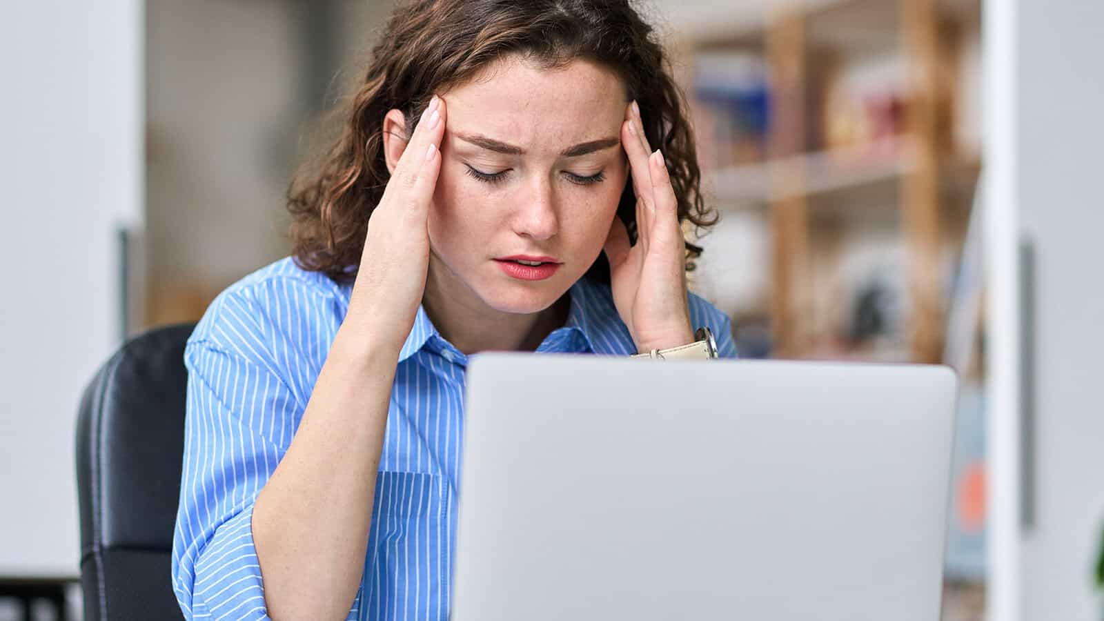 A woman sitting at a desk with her hands on her temples, looking stressed or worried while working on a laptop in a home office setting.