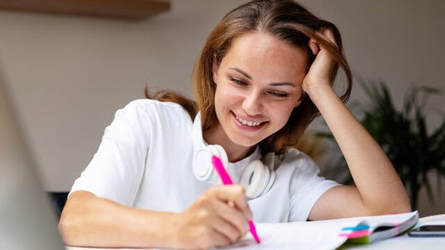 Smiling person with headphones around their neck writes with a pink pen in a notebook, appearing to study or work at a desk.