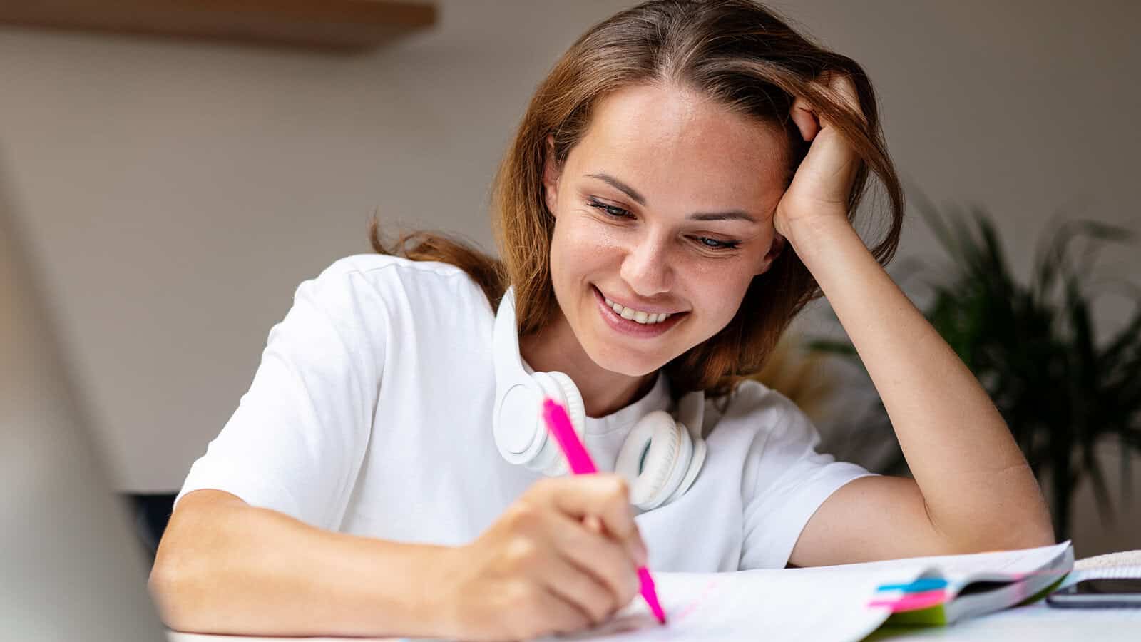 Smiling person with headphones around their neck writes with a pink pen in a notebook, appearing to study or work at a desk.