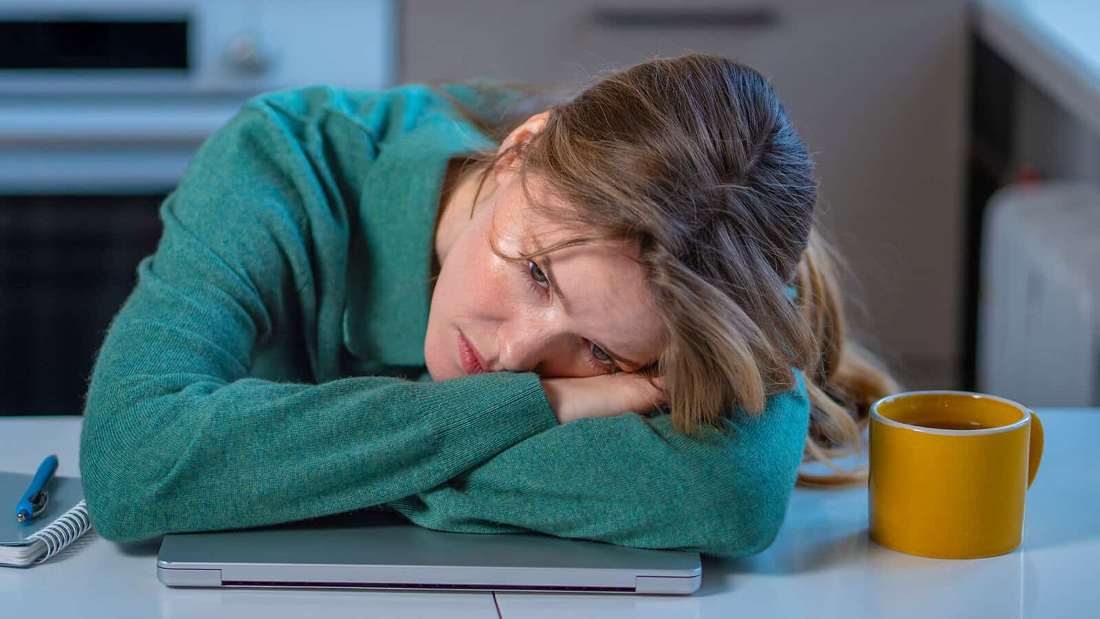 A woman in a green sweater rests her head on her folded arms atop a closed laptop, looking tired or sad. A yellow mug and a notebook with a pen are on the white table beside her.